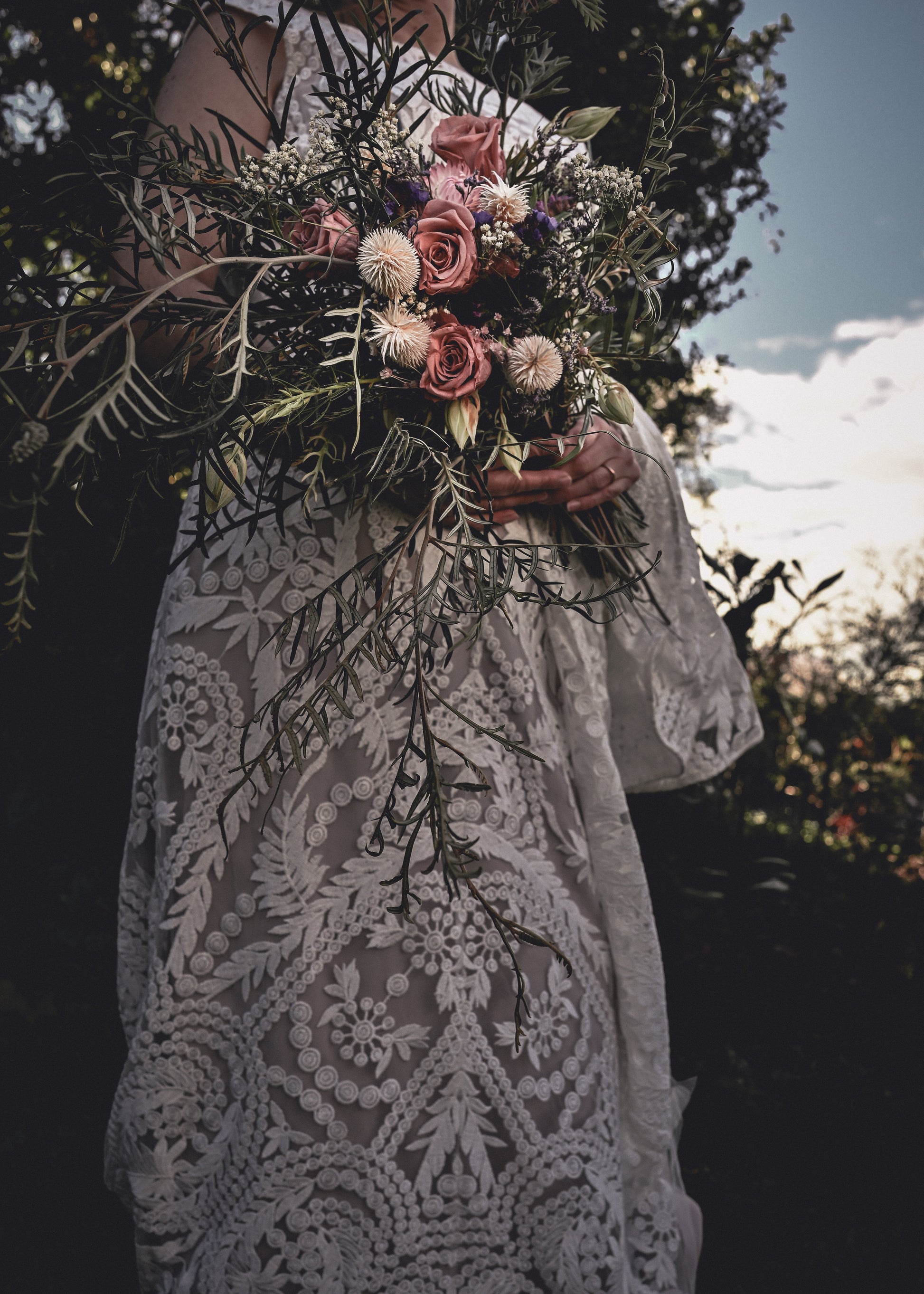 Person wearing a lace dress holding a bouquet of flowers outdoors.