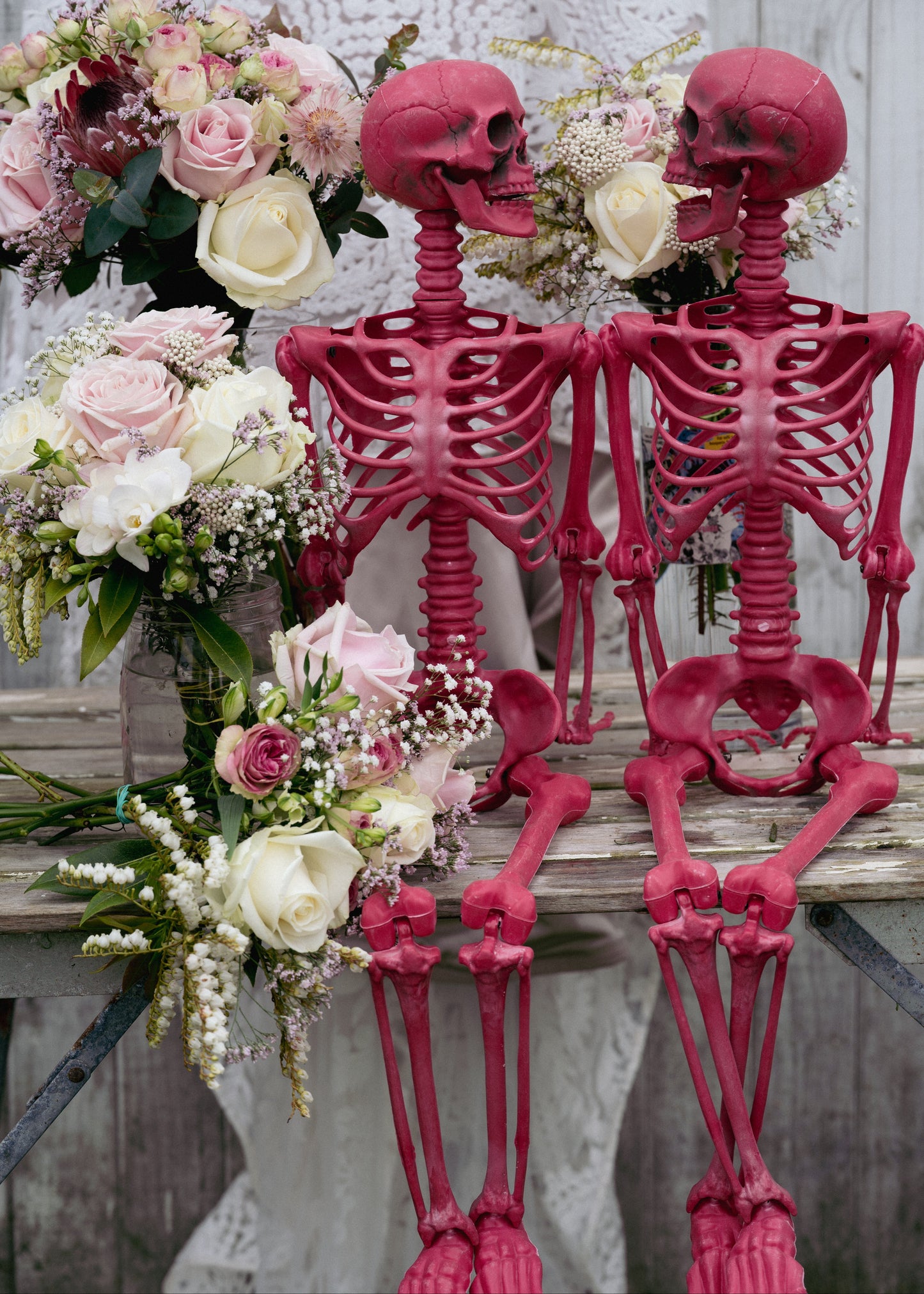 Two pink skeletons standing next to floral arrangements on a wooden surface.