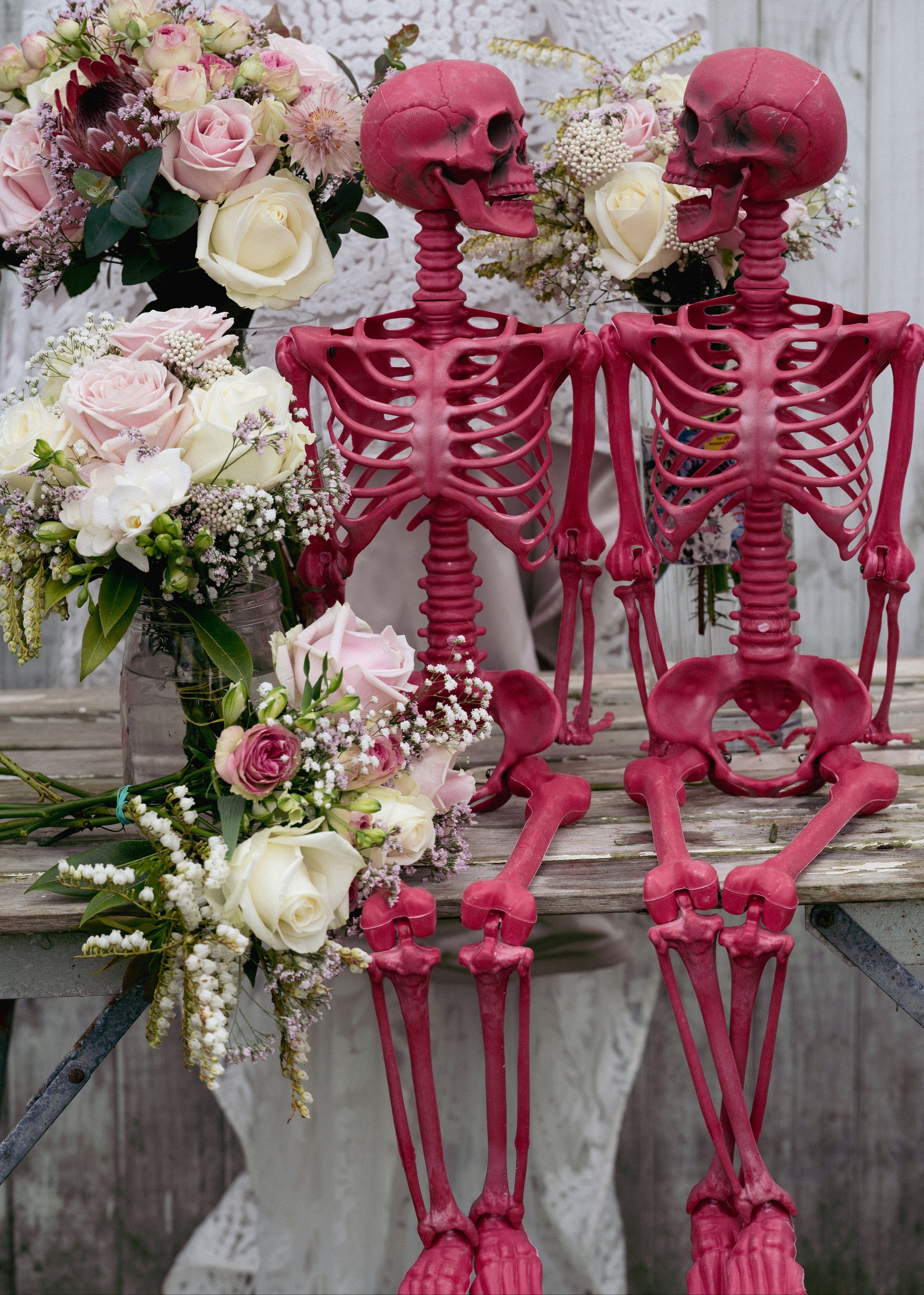 Two pink skeletons standing next to floral arrangements on a wooden surface.